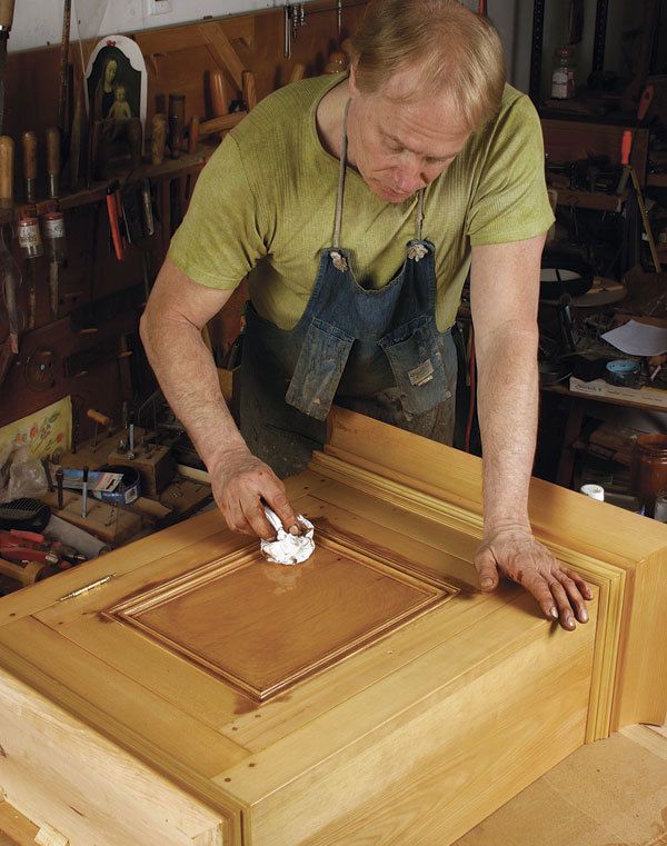 A white man rubbing darkening finish onto a golden cabinet. 